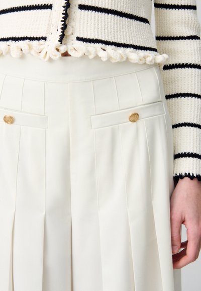 Cárdigan de punto con patrón a rayas en crema y negro; combinado con una falda blanca plisada con bolsillos laterales y detalles de botones dorados.