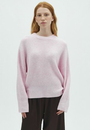 Giovane donna con capelli rossi che indossa un maglione a coste rosa chiaro e pantaloni neri larghi, in piedi davanti a uno sfondo bianco semplice.