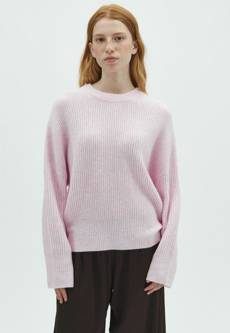 Jeune femme aux cheveux roux portant un pull côtelé rose clair et un pantalon noir ample, debout devant un fond blanc uni.