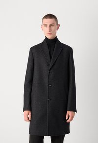 Cappotto di lana nero con finitura testurizzata, rever a doppio petto, cinque bottoni e tasche laterali. Indossato sopra un dolcevita nero.