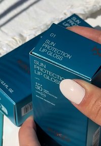 Turkos box med solskyddande läppglans, SPF 30, med vit text och röda accenter. Synlig hand med ljusfärgat nagellack.