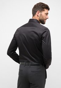 Camicia da uomo in satin nero con colletto strutturato e maniche lunghe, abbinata a pantaloni grigi scuri con una texture liscia e tasche sottili.