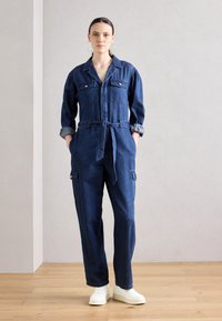 Salopette en denim avec manches longues, taille cintrée, deux poches poitrine et poches latérales de style cargo. Couleur bleu foncé, coupe décontractée.