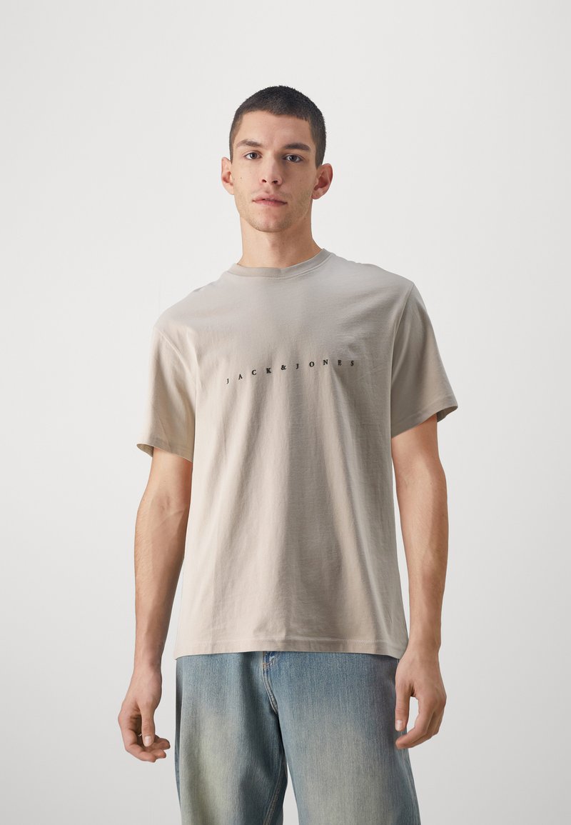 Camiseta de algodón beige de manga corta, con cuello redondo y logo de texto negro "JACK & JONES" en el pecho. Llevada con jeans de color azul claro.
