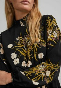 Abito nero a fiori con motivi gialli e bianchi. Presenta un colletto strutturato e arricciato e maniche lunghe con polsini elasticizzati.