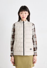 Gilet matelassé beige avec un col haut, présentant des panneaux matelassés horizontaux et une fermeture éclair à l'avant. Porté par-dessus une chemise à carreaux marron.