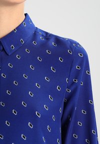 Blusa di seta blu con colletto. Il tessuto presenta un motivo geometrico ripetuto in bianco e nero. Texture liscia.