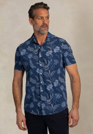 Man met een gemêleerde baard die een donkerblauw shirt met korte mouwen draagt met lichtblauwe bloemmotieven, staand tegen een effen achtergrond.