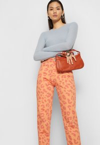Top de malha de manga longa azul claro; jeans rosa com padrão de leopardo; pequena mala laranja com ferragens em prata; mulher de pé com os braços cruzados.