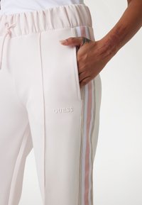 Joggers rose clair avec une finition texturée, taille élastique, poches latérales et rayures pastel sur les jambes. Avec un logo "GUESS".