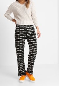 Mujer vestida con un suéter de punto color crema, pantalones negros con patrones geométricos y zapatillas de deporte naranja brillante, de pie contra un fondo blanco.