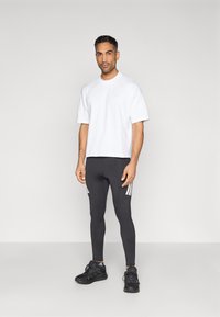 T-shirt branca de manga curta, leggings pretas com riscas brancas e tênis pretos, em pé sobre um fundo neutro.