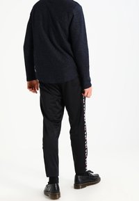 Pantalones jogger negros, de corte entallado con franjas laterales que incluyen un logo. Combinados con un suéter oscuro de punto. Textura suave y diseño casual.