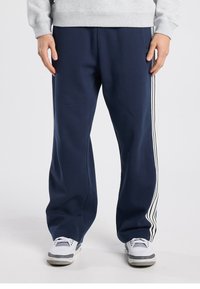 Pantalon de survêtement bleu marine en tissu doux avec des bandes latérales blanches, présentant une coupe décontractée et une taille élastique, associé à des baskets blanches.