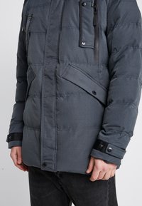 Chaqueta acolchada en gris oscuro, confeccionada con tela texturizada. Cuenta con bolsillos laterales, un bolsillo con cremallera y botones a presión en los puños para un ajuste ajustable.