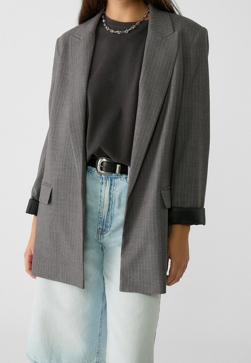 Blazer gris con rayas finas y hombros estructurados, combinado con una camisa negra y jeans de mezclilla azul claro de tiro alto. Presenta detalles en los puños de color negro.