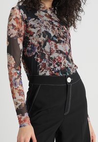 Haalimaton pitkähihainen paita, jossa on monivärinen kukkakuosi mustalla taustalla, yhdistettynä korkeavyötäröisiin mustiin housuihin, joissa on valkoiset saumat.