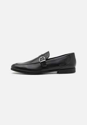 Mocassin en cuir noir à enfiler avec un détail de boucle argentée et un petit talon, embossé sur le côté du logo de la marque.