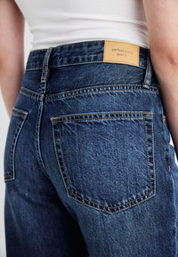 Dark blue denim jeans with a high waist, visible stitching, two back pockets, and a brown leather patch labeled "perfect jeans."