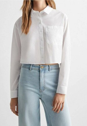 Frau trägt ein weißes, kurz geschnittenes Langarmhemd mit Knöpfen, einer Brusttasche und hellblauen, hoch taillierten Jeans.