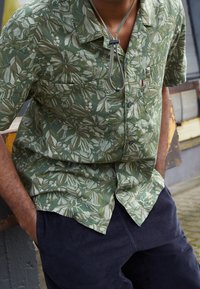Camicia verde a maniche corte con motivo floreale in tonalità di verde e beige, caratterizzata da una tasca sul petto e colletto con bottoni.