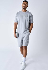 Ensemble en coton gris comprenant un t-shirt et un short avec logo brodé. Le mannequin tient une petite pochette grise. Des baskets blanches et des chaussettes complètent la tenue.