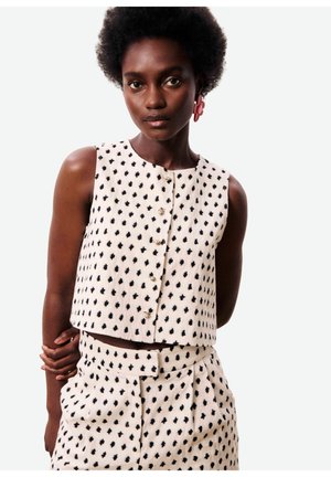 Femme aux cheveux courts naturels portant un haut crème sans manches et une jupe taille haute assortie avec un motif de points noirs abstraits, et des boucles d'oreilles en forme de fleurs roses.