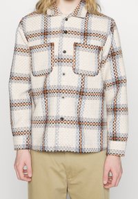 Vävd rutmönstrad skjorta i beige, grå och orange, med två bröstfickor och en strukturerad tyg. Slät krage och svarta knappar.