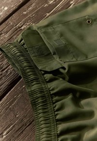 Közelkép olívazöld nadrágról, rugalmas derékkal és tépőzárral zárható hajtóka zsebbel, textúrált barna fa felületen.