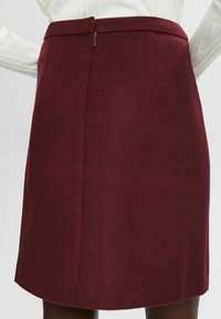Gros plan d'une personne portant une jupe en laine bordeaux taille haute, longueur genou, avec une fermeture éclair au dos et un pull blanc côtelé.