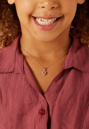 Gold-tone necklace with a heart-shaped pendant featuring a central pink stone, worn over a textured, maroon button-up shirt.