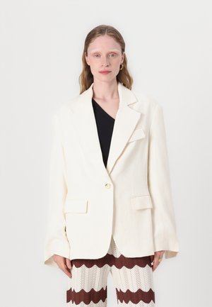 Cream-colored blazer with a notched lapel, single button closure, and two front pockets, paired with patterned knit pants.