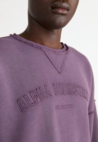 Lilla sweatshirt med brodert "ALPHA INDUSTRIES" tekst og "EST. SINCE 1959," med rund hals og rå kantdetaljer.