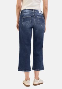 Jeans en denim bleu foncé, cropped, avec une jambe droite, dotés de deux poches arrière et d'une finition légèrement usée, associés à des baskets beiges.