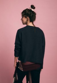 Dunkelgrauer übergroßer Pullover mit Rundhalsausschnitt und Raglanärmeln, kombiniert mit einem geschichteten rot-burgunderfarbenen Oberteil und dunklen Jeans.