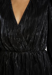 Svart plisserad klänning med djup v-ringning, figursydd midja och glänsande, texturerad tyg som skapar en svävande look.