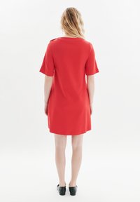 Kurzes rotes Kleid mit kurzen Ärmeln und einem Boot-Ausschnitt, glatter Textur und einer lockeren Passform, das oberhalb der Knie endet. Es werden schwarze flache Schuhe getragen.