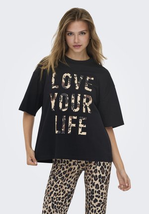 Joven mujer con camiseta negra holgada con el texto "AMA TU VIDA" y pantalones de estampado de leopardo, posando contra un fondo liso.