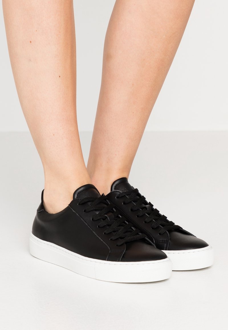 Pieds chaussés de baskets basses en cuir noir avec lacets noirs et semelles en caoutchouc blanches épaisses, sur un fond clair.