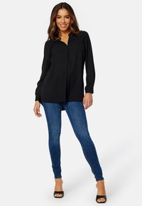 Blusa nera a maniche lunghe con bottoni sulla parte anteriore, abbinata a jeans skinny blu e tacchi aperti neri. Tessuto liscio e vestibilità su misura.