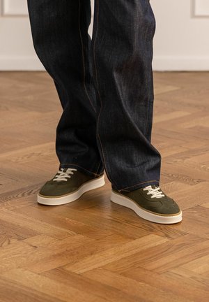 Persona che indossa jeans blu scuro e sneakers verde oliva con suola bianca, in piedi su un pavimento in parquet di legno.