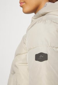 Lys beige hættetrøje pufferjakke med en struktureret overflade, der har et sort stoflogo-patch på venstre ærme.
