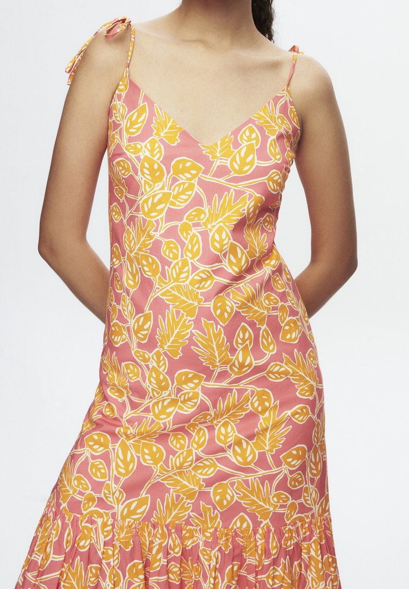 Femme portant une robe rose avec des motifs de feuilles jaunes et des fines bretelles nouées aux épaules, debout les mains dans le dos.