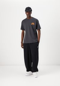 Mørkegrå Nike T-shirt med orange logo, sammen med løse sorte bukser. Hvide sneakers med et tekstureret mønster og en sort kasket. Minimalistisk design.