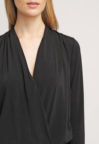 Blouse noire à enrouler avec un décolleté en V profond, des manches longues et un tissu doux et lisse avec un léger brillant. Design simple, sans motifs ni embellissements.