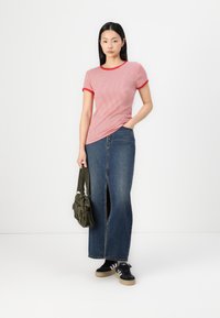 Gestreiftes, eng anliegendes T-Shirt in Rot und Weiß mit kontrastierendem roten Saum, kombiniert mit einem langen blauen Jeansrock mit einem Schlitz vorne. Accessoires: eine grüne Tasche.