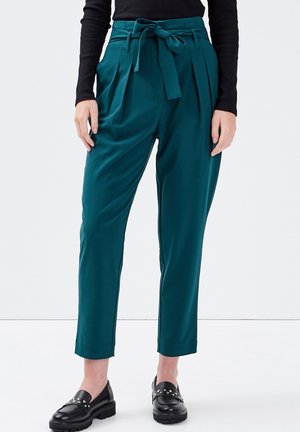 Pantalon taille haute couleur sarcelle avec plis à l'avant et ceinture nouée, porté avec un haut à manches longues noir et des mocassins noirs cloutés.
