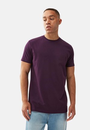 Kortærmet, rundhalset T-shirt i dyb lilla bomuldsstof, parret med lyseblå denimjeans. Har et subtilt logo ved kanten.