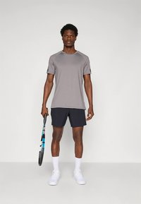 Grijs sportief T-shirt met korte mouwen, gecombineerd met donkere shorts en witte sokken. Het model houdt een blauw-zwarte tennisracket vast.