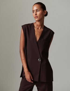 Γυναίκα με τα μαλλιά τραβηγμένα προς τα πίσω που φοράει ένα αμάνικο σκούρο καφέ σακάκι με ένα ασημί κουμπί, στέκεται μπροστά σε μια απλή γκρι φόντο.